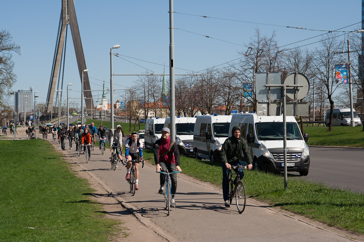 A group of people are riding bicycles along a path on a clear day. The path runs parallel to a road where several white vans are parked. A tall bridge structure with cables is visible in the background. Leafless trees line the grassy area next to the path, suggesting early spring reminiscent of Kritiskā masa 2012.
