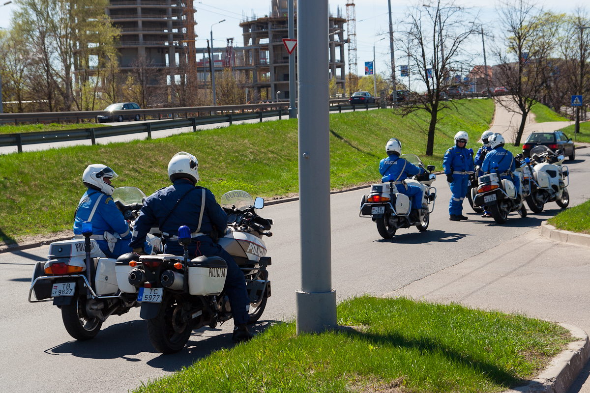 Four police officers on motorcycles, dressed in blue uniforms and white helmets, patrol an urban road during the Kritiskā masa 2012 event. Two officers face forward on the left side, while the other two face backward on the right. A construction site and sparse trees are visible in the background. Green grass lines the road.