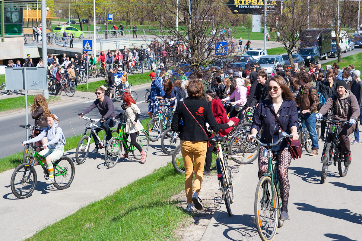 A large group of people participating in the 2012 Kritiskā masa bike ride event on a sunny day. Men, women, and children walk with and ride bicycles on a sidewalk and grass area along a busy road. Some participants are smiling, and there are trees and buildings visible in the background.