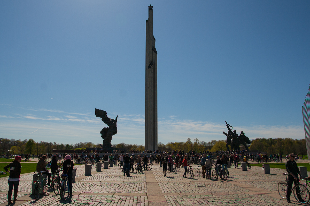A large outdoor plaza bathed in sunlight features two prominent statues on either side and a tall central monument. Numerous people with bicycles, reminiscent of Kritiskā masa 2012, are gathered, enjoying the day under a clear blue sky. Trees in the background are slightly autumnal, offering a splash of green and yellow hues.