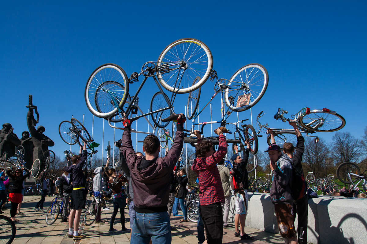 A group of people are gathered outdoors on a sunny day, raising their bicycles above their heads in a celebratory manner. The sky is clear and blue, and people are dressed in casual clothing. Some individuals are seen smiling and interacting with each other, reminiscent of the Kritiskā masa event in 2012.