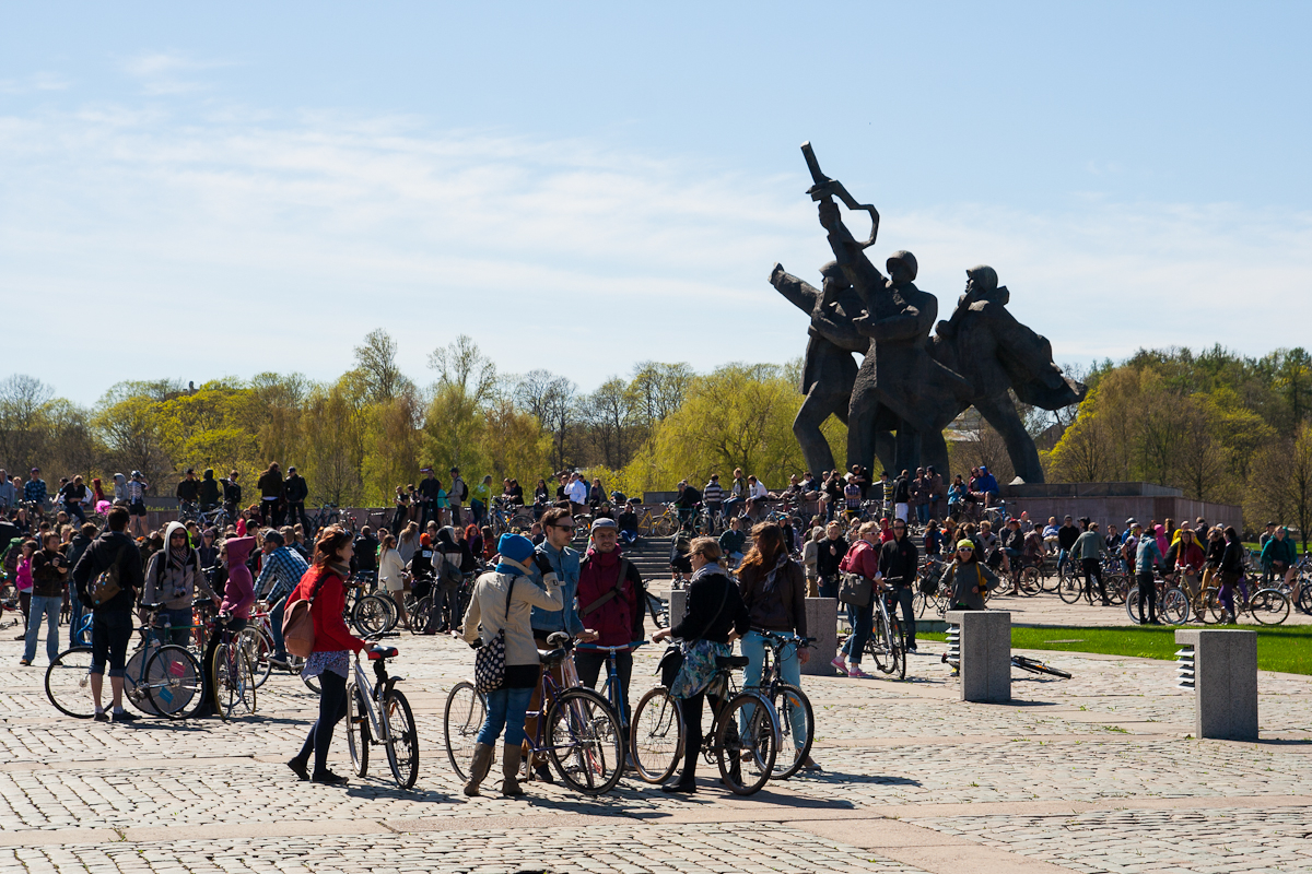 A large group of people gather on a cobblestone square with bicycles, reminiscent of the 2012 Kritiskā masa event. In the background, there's a tall, abstract sculpture of several figures. Trees with light green foliage mark the horizon under a clear blue sky. The relaxed crowd is engaged in conversations and activities.