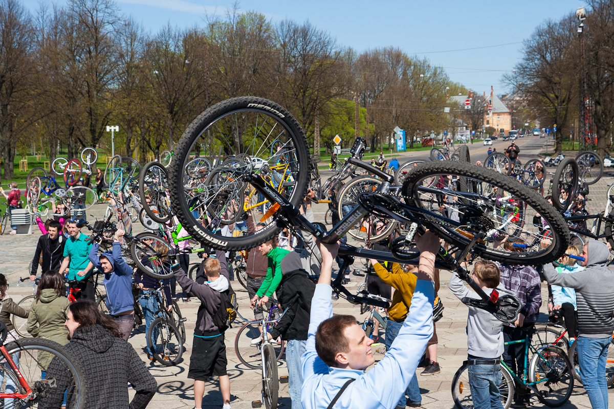 A large group of people gathered outdoors in a park on a sunny day, many holding up bicycles over their heads. The crowd is diverse, and trees in the background are budding, suggesting springtime. This 2012 event appears to be Kritiskā masa, celebrating or demonstrating the joys of cycling.