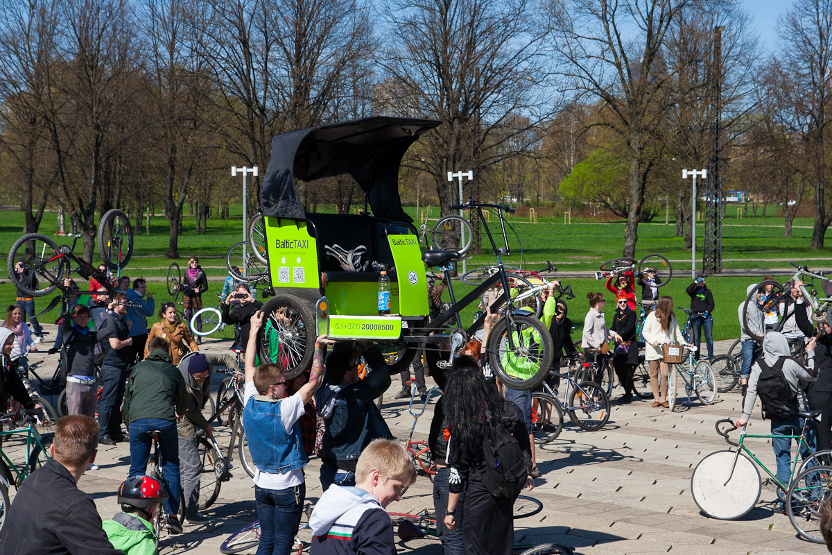 A lively scene of a large group of people gathering outdoors in a park for the 2012 Kritiskā masa event. Some are holding up their bicycles, and a green pedicab with a black canopy is prominently displayed in the center. The weather appears sunny, and the background features leafless trees and green grass.