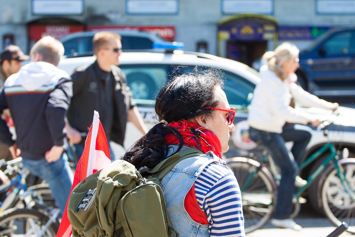 A person with a black ponytail, wearing red sunglasses, a striped shirt, and a green backpack with a red-and-white flag attached, is seen from behind during the 2012 Kritiskā masa. They are part of a group of cyclists. Several people on bicycles and a police car are in the background on a sunny day.