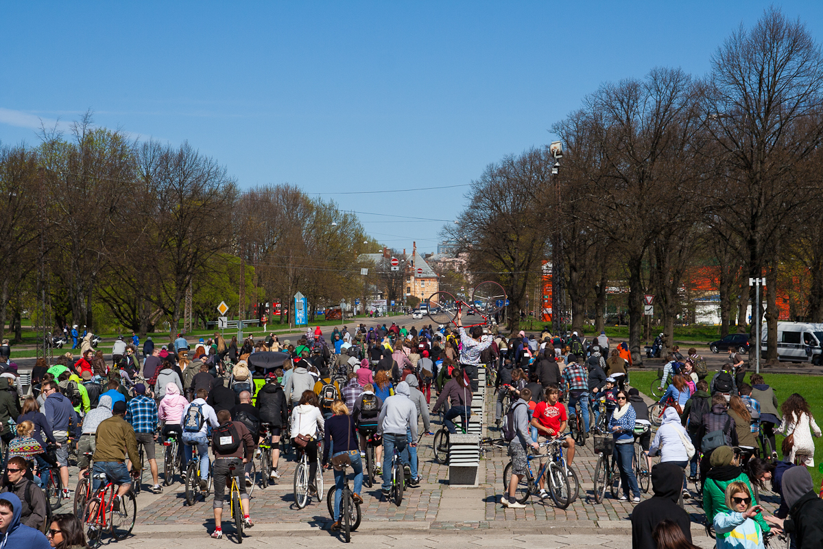 A large crowd of cyclists and pedestrians, viewed from behind, participate in the 2012 Kritiskā masa event on a sunny day. The group is moving along a broad pathway lined with leafless trees. Participants wear casual attire, and some carry backpacks. Blue skies and distant buildings are visible in the background.