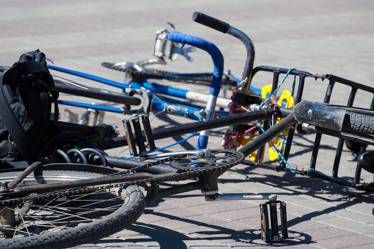 Two bicycles lie on the ground on a paved surface during Kritiskā masa 2012. One has a blue frame with black handlebar tape and no visible front wheel, while the other has a black frame with the pedal and gear area prominently showing. A black backpack is partially visible near the bicycles.