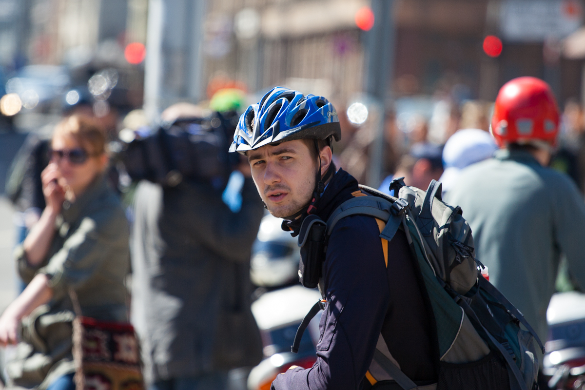 A cyclist wearing a blue helmet, black long-sleeve shirt, and a backpack pauses amidst a busy city street during Kritiskā masa 2012. He looks ahead while people around him, some also on bikes, engage in various activities. The background is blurred, showing a mix of pedestrians and urban elements.