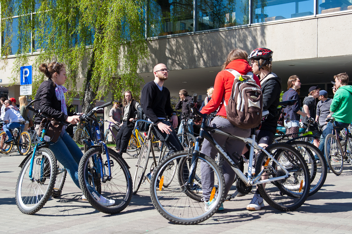 A group of people with bicycles gather outside under a sunny sky. The scene, reminiscent of Kritiskā masa 2012, includes both men and women dressed in casual attire, conversing animatedly. A modern building with large windows and a tree with green leaves form the background, creating a lively and relaxed atmosphere.