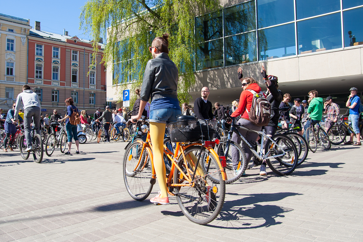 A group of people with bicycles gather in a sunny urban plaza near a modern glass building. One person with orange tights and a denim jacket stands prominently in the foreground holding a yellow bicycle. The crowd, reminiscent of the 2012 Kritiskā masa event, appears relaxed, some standing and talking, others seated on their bikes.