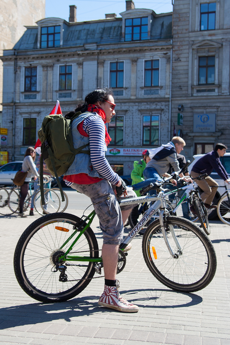 A cyclist stands beside their bicycle on a city street, preparing for a ride. They are wearing a striped long-sleeve shirt, camouflage shorts, and red sneakers with white stars. A green backpack and a red flag are attached to the bike. Other cyclists and historic buildings are visible in the background, reminiscent of Kritiskā masa 2012.