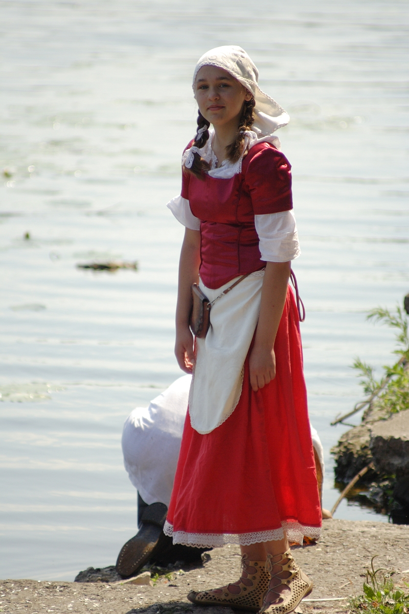 A young woman stands by a lake, dressed in a red historical costume with a white apron and headscarf. She has braided hair and wears lace-up shoes. The calm waters of the Dunamunde shoreline provide a serene backdrop, while another person kneels near the water, mostly out of view.