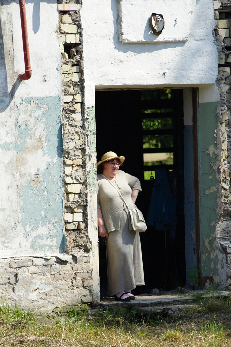 A person wearing a beige outfit and a straw hat stands in the doorway of a partially dilapidated Dunamunde 2010 building with peeling paint and exposed bricks. They have a white bag across their shoulder and are looking out into the sunlight. The area around the building appears grassy and untended.