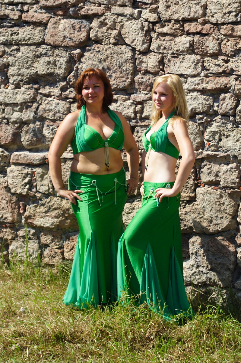 Two women stand in front of a stone wall, wearing matching green belly dance costumes, reminiscent of Dunamunde 2010. The woman on the left has short red hair, and the woman on the right has long blonde hair. Both are smiling and posing with one hand on their hip. Grass is visible at the bottom of the image.