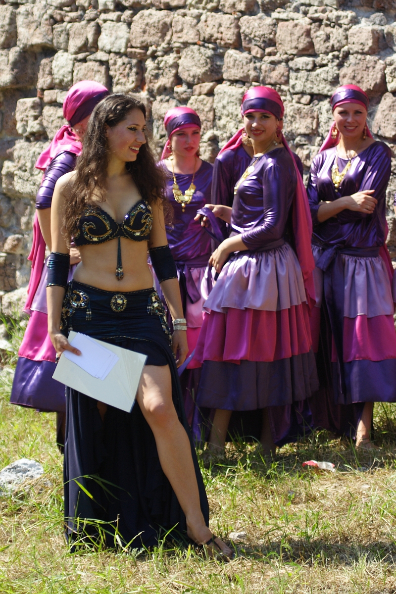 A woman with long, dark hair and a belly dance costume stands in the foreground, holding a paper. Her costume includes a black and gold top and flowing black skirt. Behind her, several women in purple and pink dresses and headscarves stand in a group near a stone wall, reminiscent of Dunamunde performances from 2010.