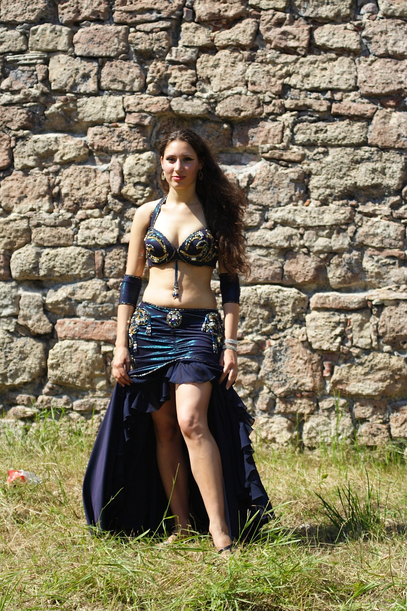 A woman with long curly hair stands in front of a stone wall, reminiscent of Dunamunde 2010. She is wearing a dark blue belly dance costume adorned with gold embellishments. The outfit includes a top, arm bands, and a skirt with a high slit, revealing part of her leg. Grass is visible at her feet.