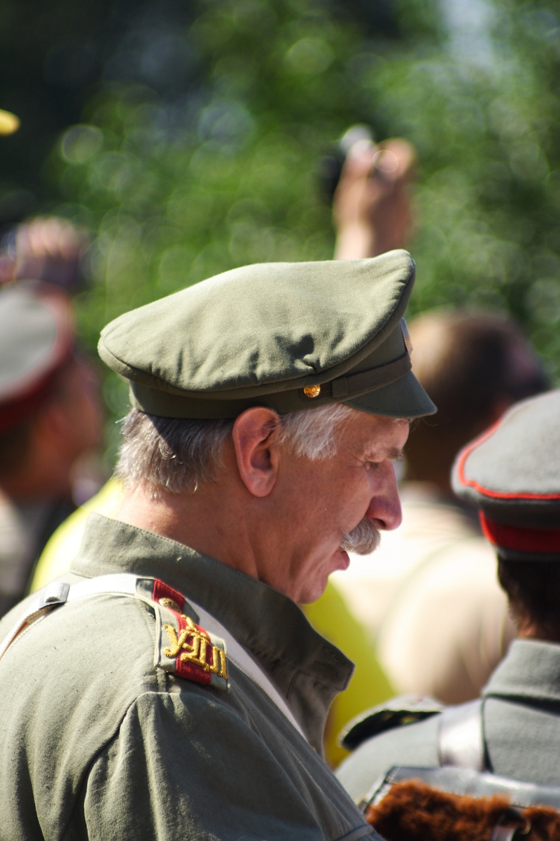 A man with a gray mustache wearing a vintage military uniform and a matching cap stands outdoors with his head slightly bowed at the Dunamunde 2010 event. The uniform features red and gold shoulder epaulettes. Blurred figures in similar attire are visible in the background, suggesting a group or gathering.