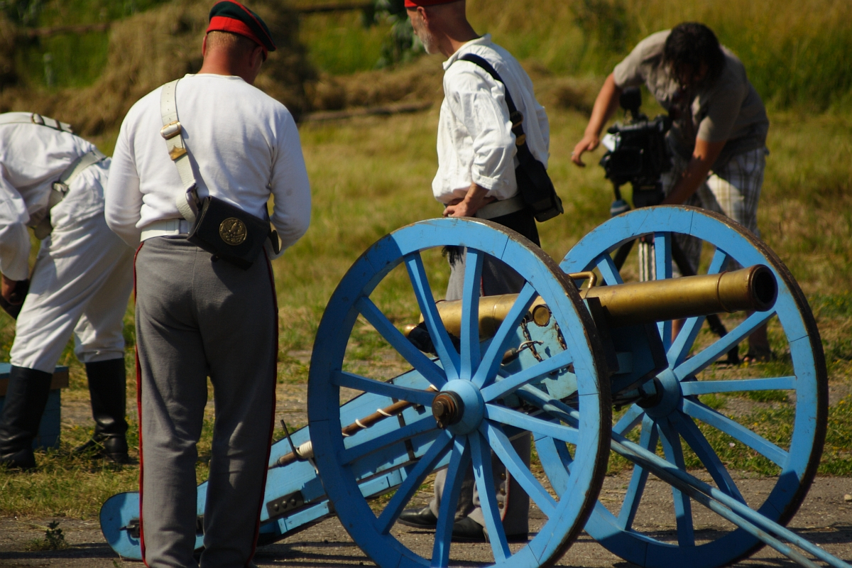 Several people in historical military attire stand around a bright blue cannon with large wheels in an outdoor setting. Some are adjusting the cannon, while another person in modern clothing appears to be filming the scene, likely capturing the 2010 Dunamunde reenactment. Dry grass and stacked hay are visible in the background.