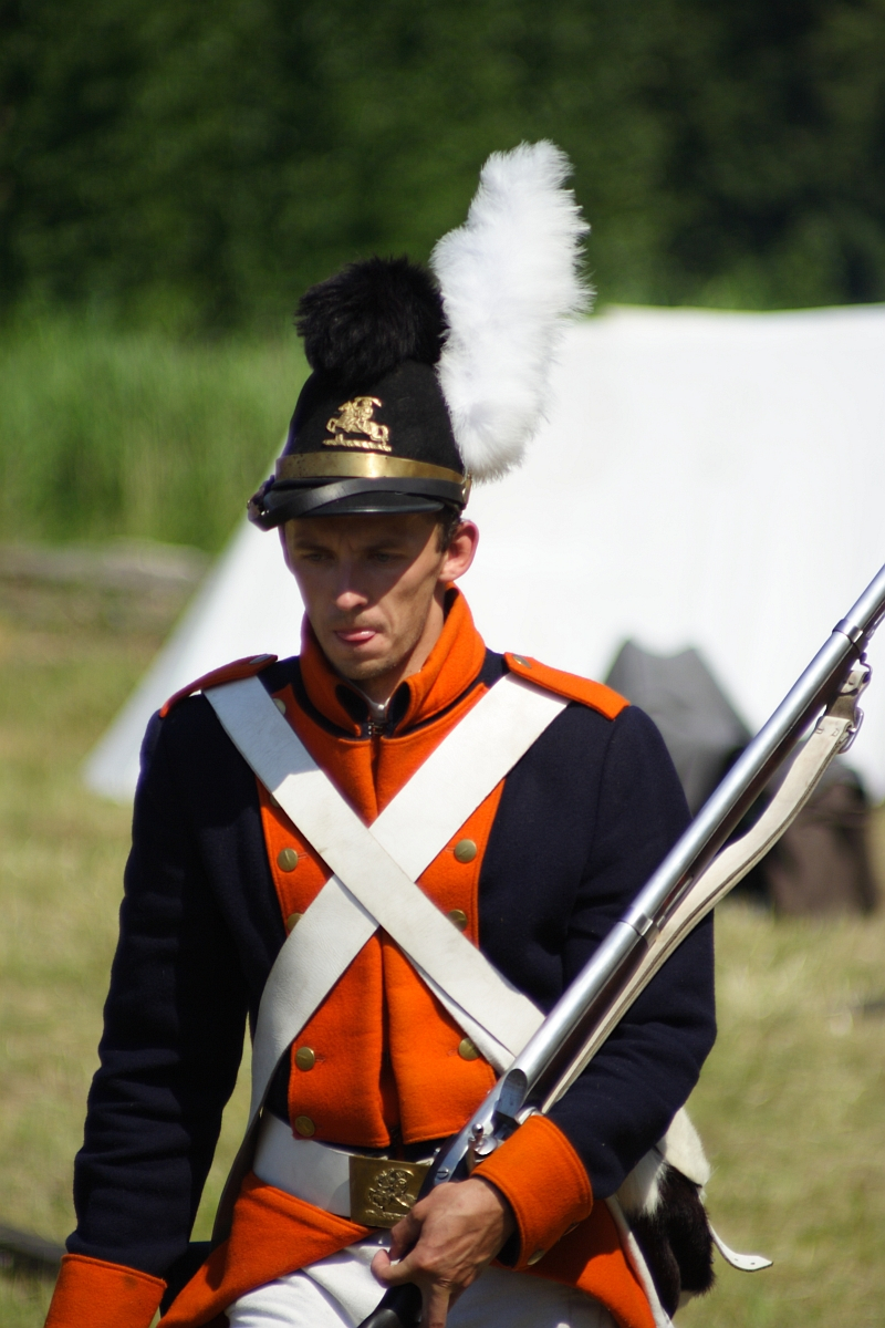 A man dressed as a historical soldier stands outdoors at Dunamunde 2010, holding a rifle with a bayonet. He's wearing a navy blue and orange uniform with white crossbelts, a tall shako hat adorned with a black and white plume, and brass insignia. The background features blurred greenery and a white tent.