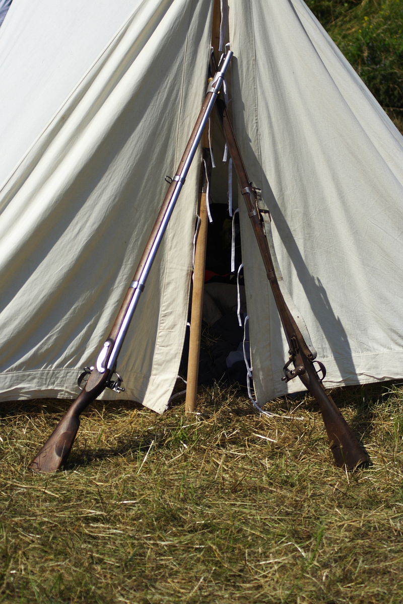 A canvas tent is set up on a grassy area, with its entrance open. Two rifles, reminiscent of reenactments like Dunamunde 2010, are propped up against the tent, crossing each other at the entrance. The ground around the tent is grassy, showcasing a rustic outdoor setting. The interior of the tent is not visible.