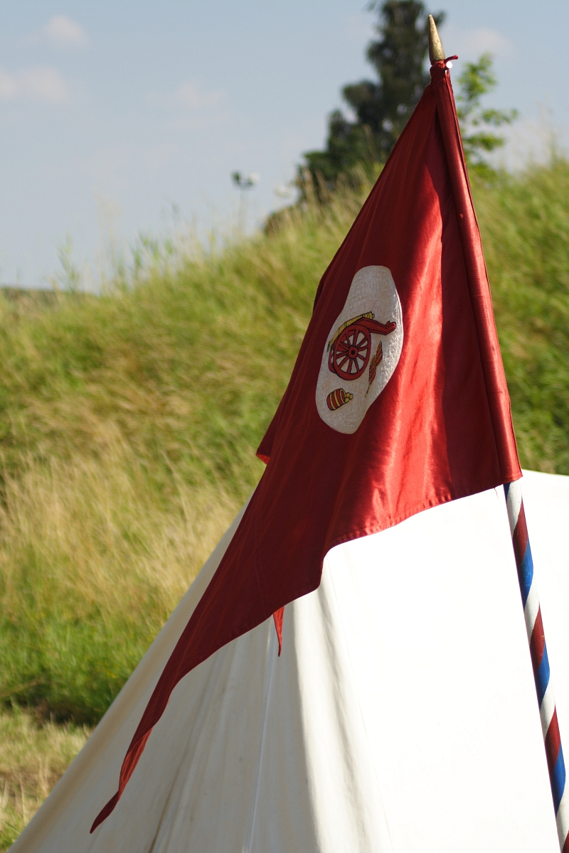 A red flag, adorned with a white badge featuring a medieval knight's helmet and crossed anchors, is mounted on a striped pole. This scene evokes the 2010 Dunamunde festival, set against a backdrop of green vegetation and a clear sky in front of a white tent pitched in a grassy area.