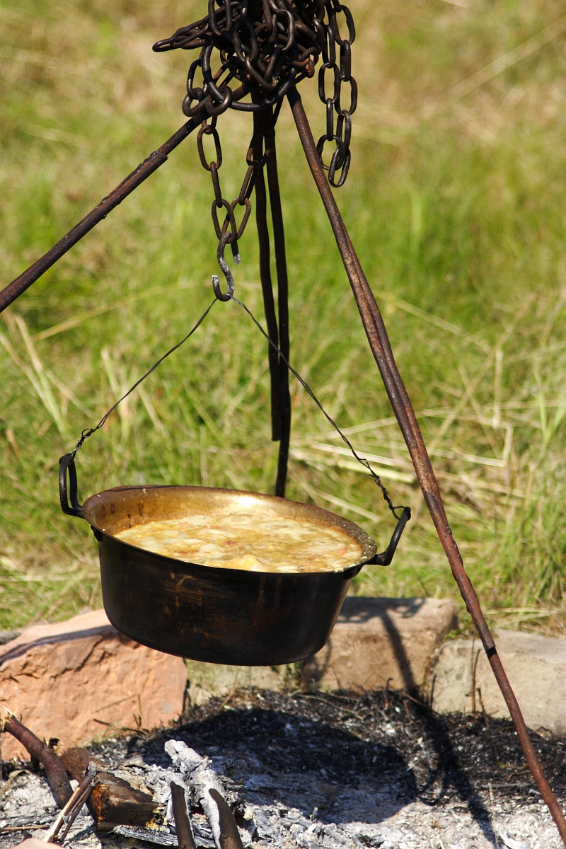A cast-iron pot filled with food hangs over a fire pit, reminiscent of Dunamunde 2010. The pot is suspended by chains attached to a metal tripod, while ashes and small burned wood pieces scatter below. The grassy terrain in the background sets the scene for outdoor cooking or camping.