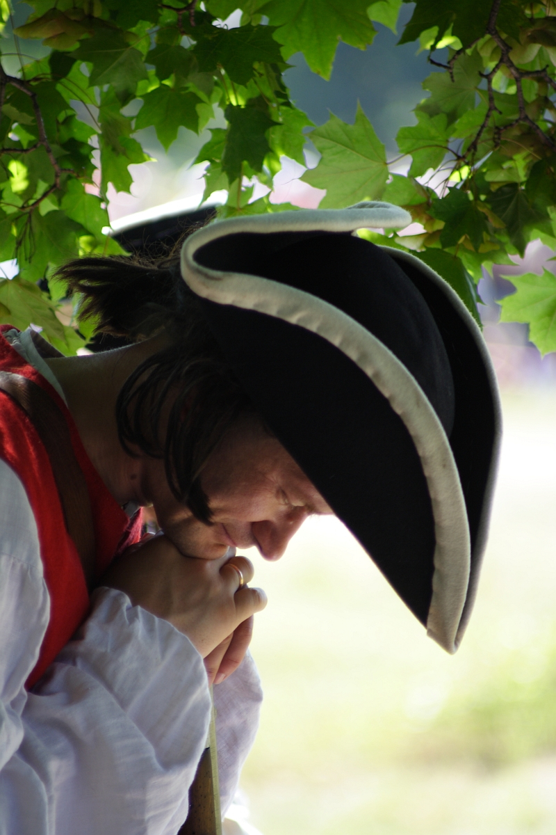 A person dressed in historical attire is depicted, wearing a tricorn hat with white trim, a white shirt, and a red vest reminiscent of the Dunamunde 2010 reenactment. They are under a leafy tree, looking down while holding a long wooden object close to their face. The scene is outdoors with a blurred background.