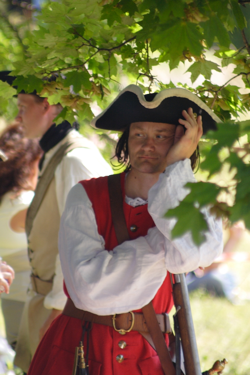 A man wearing a tricorn hat and 18th-century-style red uniform with white sleeves rests against a leafy tree branch. He has a serious expression and holds his head with one hand. Part of the Dunamunde 2010 reenactment, another person in similar historical attire is blurred in the background among green foliage.