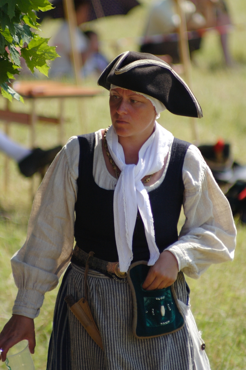 A person dressed in historical attire stands outdoors. They wear a black tricorne hat, a white headscarf, a dark vest over a beige long-sleeve blouse, and a striped apron. Holding a green pouch, they appear to be part of the Dunamunde 2010 reenactment, with blurred people and tents in the background.