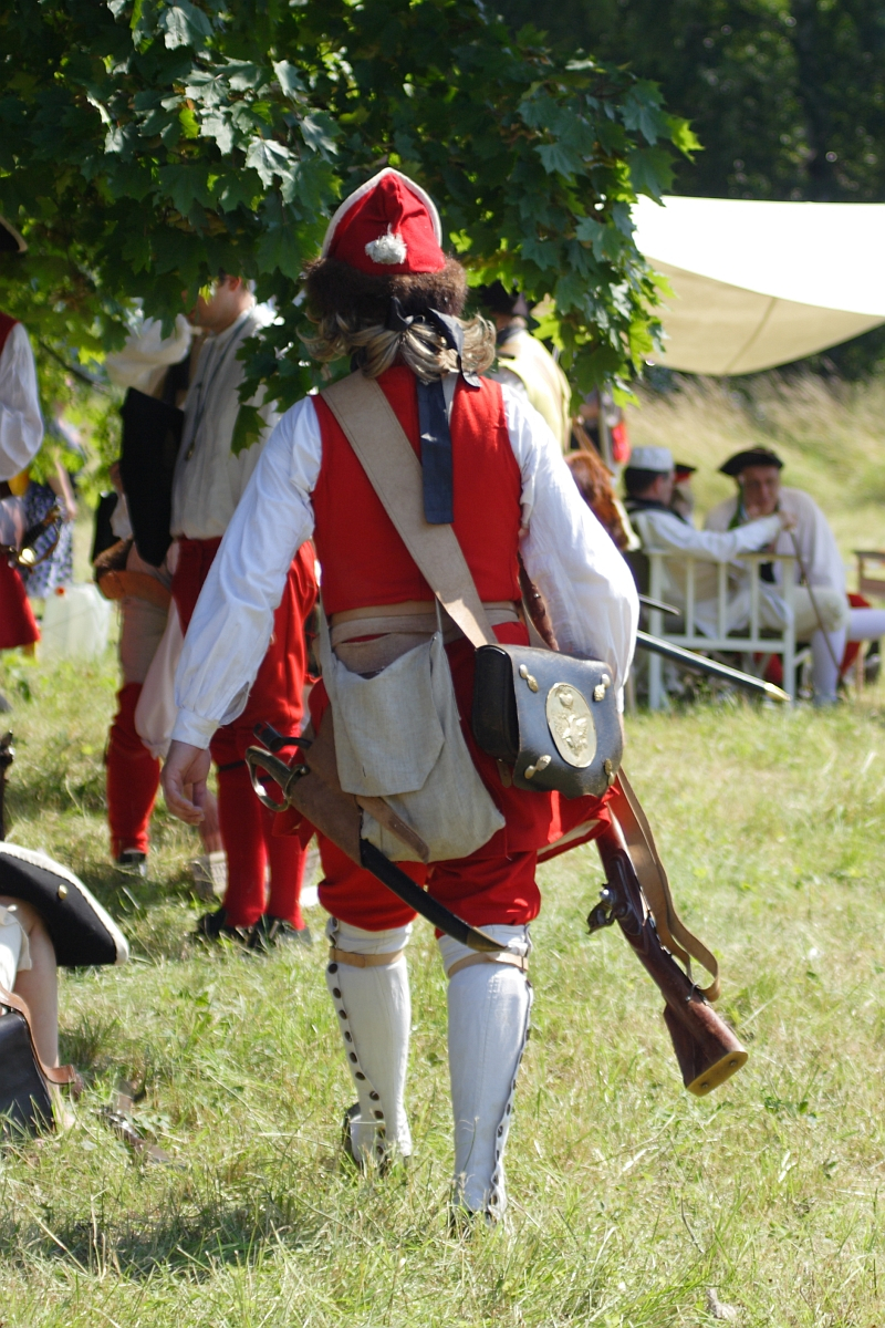A person dressed in historical military attire, reminiscent of Dunamunde 2010, including a red coat, white pants, tall boots, and a red hat, walks away from the camera on a grassy field. The person carries a musket and a satchel while other similarly dressed individuals are seated in the background under a tree.