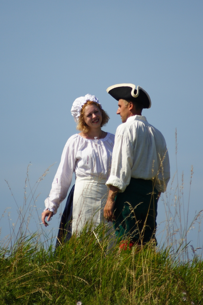 Two people dressed in historical costumes are standing in a grassy field under a clear blue sky. The woman, smiling, is wearing a white bonnet, blouse, and apron, while the man is in a white shirt and tricorn hat. They appear to be reenacting an event from Dunamunde in 2010 as they engage in conversation.