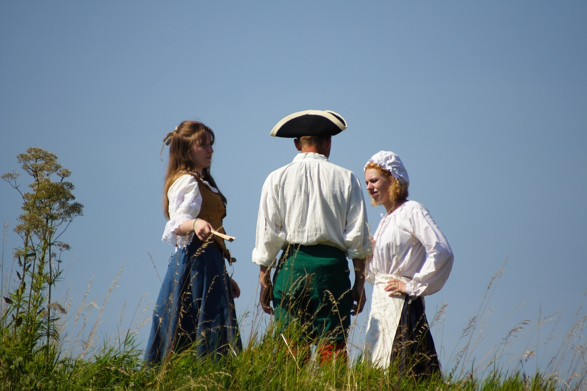 Three people dressed in historical clothing stand on a grassy hill against a clear blue sky. One woman in a blue skirt and brown vest holds a scroll, while another in an apron and bonnet interacts with them. A man in a tricorn hat and green pants stands between the two women. Tall grass and wildflowers surround them, evoking the spirit of Dunamunde despite the year being 2010