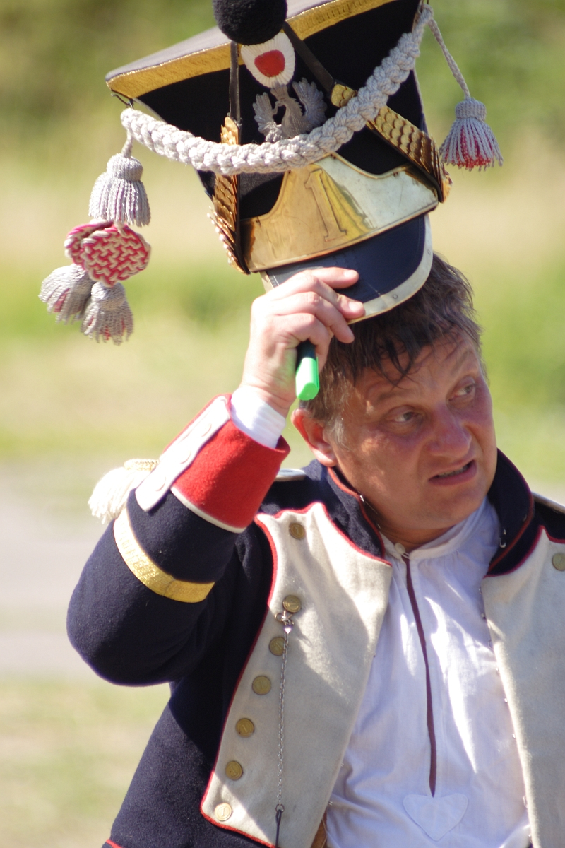 A man dressed in a historical Dunamunde military uniform, featuring a dark jacket with red and white cuffs, gold buttons, and a cream-colored vest. He is lifting a tall, ornate hat with gold embellishments, tassels, and the number "1" on it. The background is blurred, showing outdoor greenery.