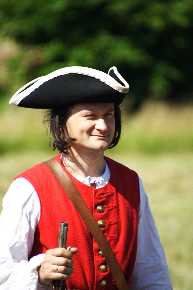A man in historical attire stands outdoors on a sunny day, wearing a black tricorn hat with white trim, a red waistcoat with brass buttons over a white long-sleeved shirt, and holding a musket. The background is blurred with greenery. He is smiling and looking into the distance, reminiscent of Dunamunde reenactments from 2010.