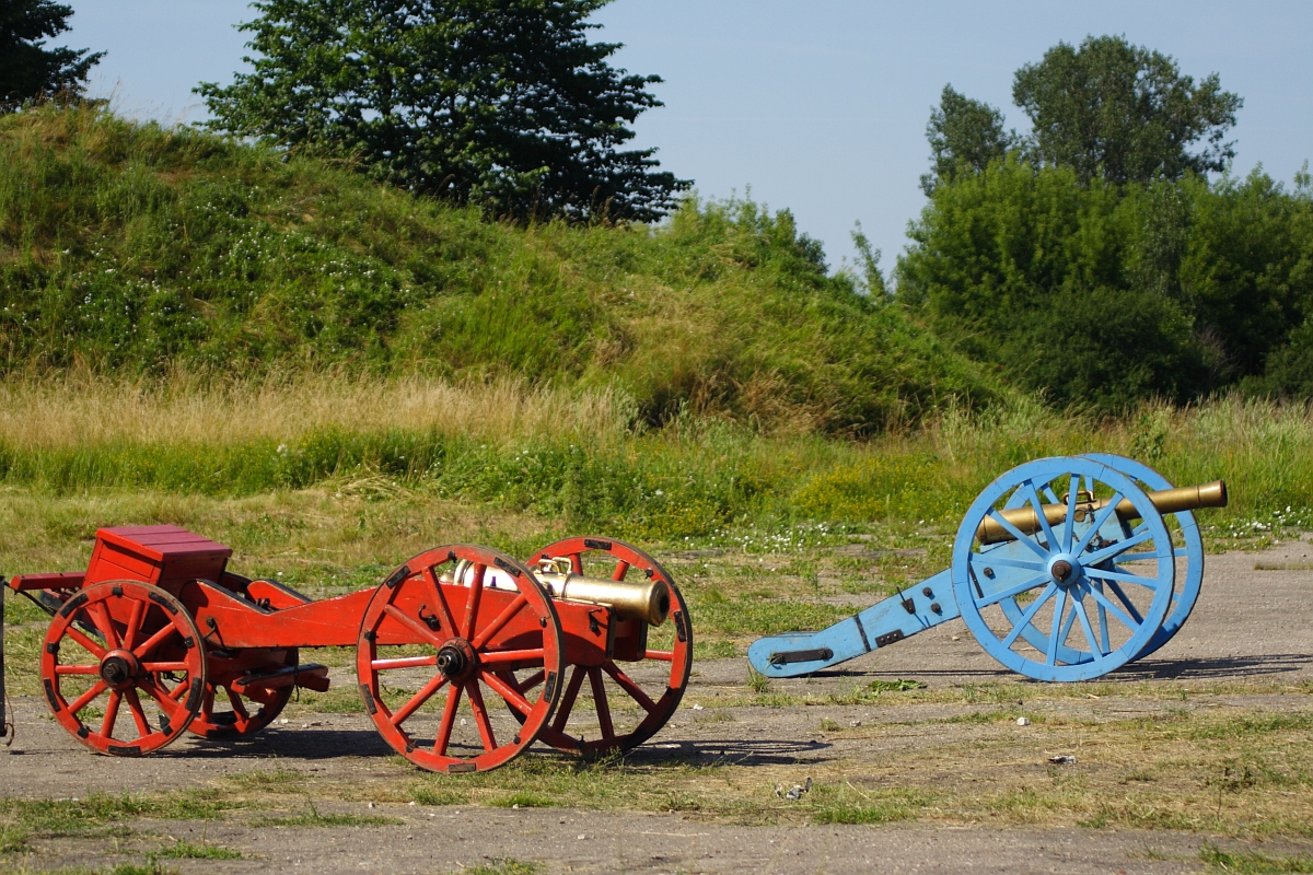 Two colorful cannons are situated outdoors on a sunny day. The cannon on the left is painted red with yellow accents, while the one on the right is blue with golden details. They are displayed on a paved surface with lush, grassy mounds and trees in the background, reminiscent of Dunamunde 2010.