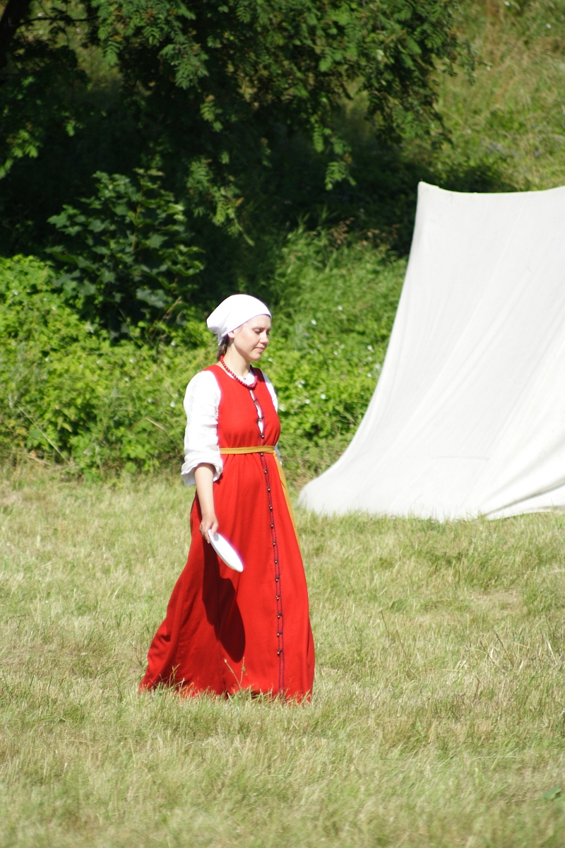 A woman in historical costume walks on grass at what appears to be a 2010 reenactment event. She wears a bright red dress with long sleeves and a white headscarf, holding a folded white paper in her right hand. Behind her, greenery and a white cloth setup, possibly a tent, completes the scene reminiscent of Dunamunde.