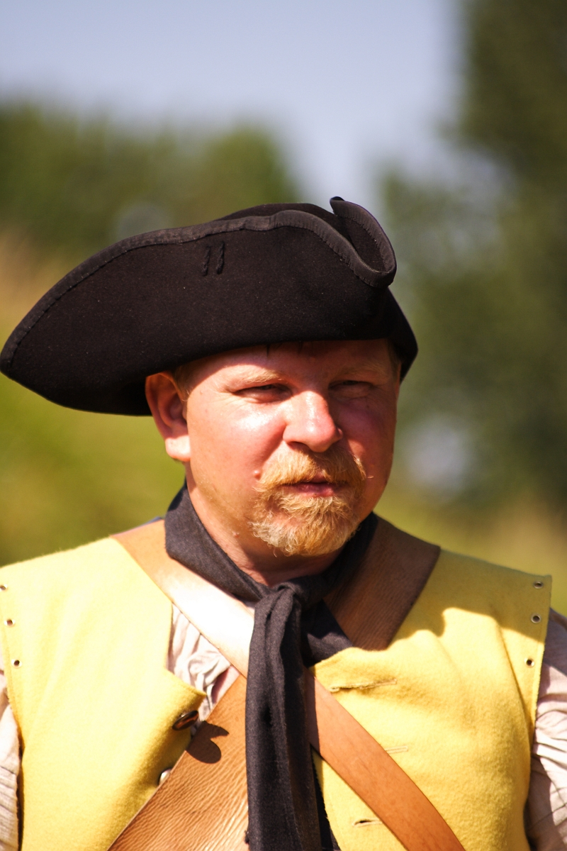 A man dressed as a historical reenactor for Dunamunde 2010 wears a tricorne hat, a yellow vest over a white shirt, and a brown leather strap across his chest. He has a light beard and stands against an outdoor background blurred with greenery. The expression on his face is serious and focused.