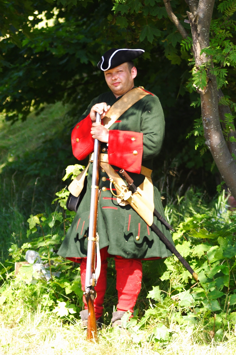A man dressed in historical military attire stands in a lush, green setting, reminiscent of Dunamunde 2010. He wears a dark green coat with red cuffs and trim, red pants, and a tricorn hat. Holding a long rifle with a beige strap across his chest, the background features green foliage and a partially visible tree trunk.