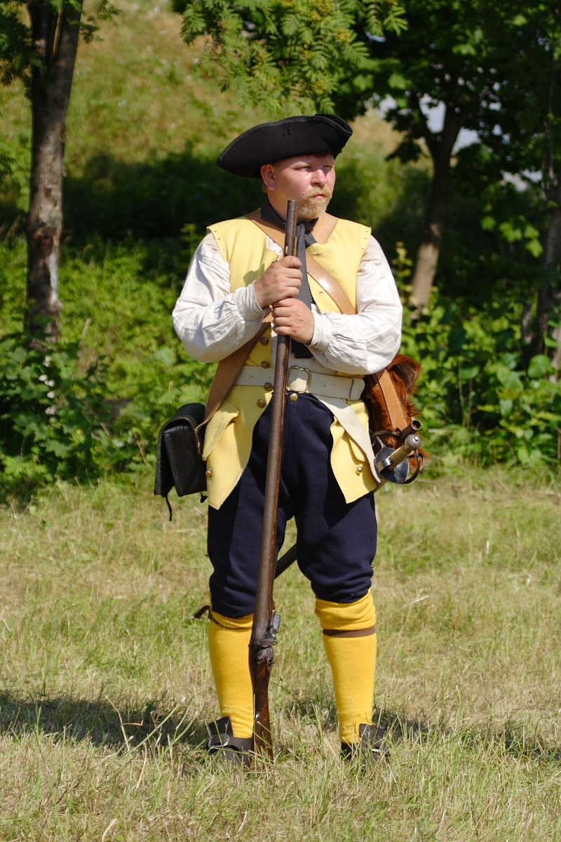 A man dressed in colonial-era clothing stands in a grassy field at the Dunamunde reenactment. He wears a tricorn hat, a light-colored vest over a white shirt, dark blue trousers, and yellow socks. He holds a long rifle upright in front of him and has various pouches and equipment strapped to his waist. Trees form the backdrop.