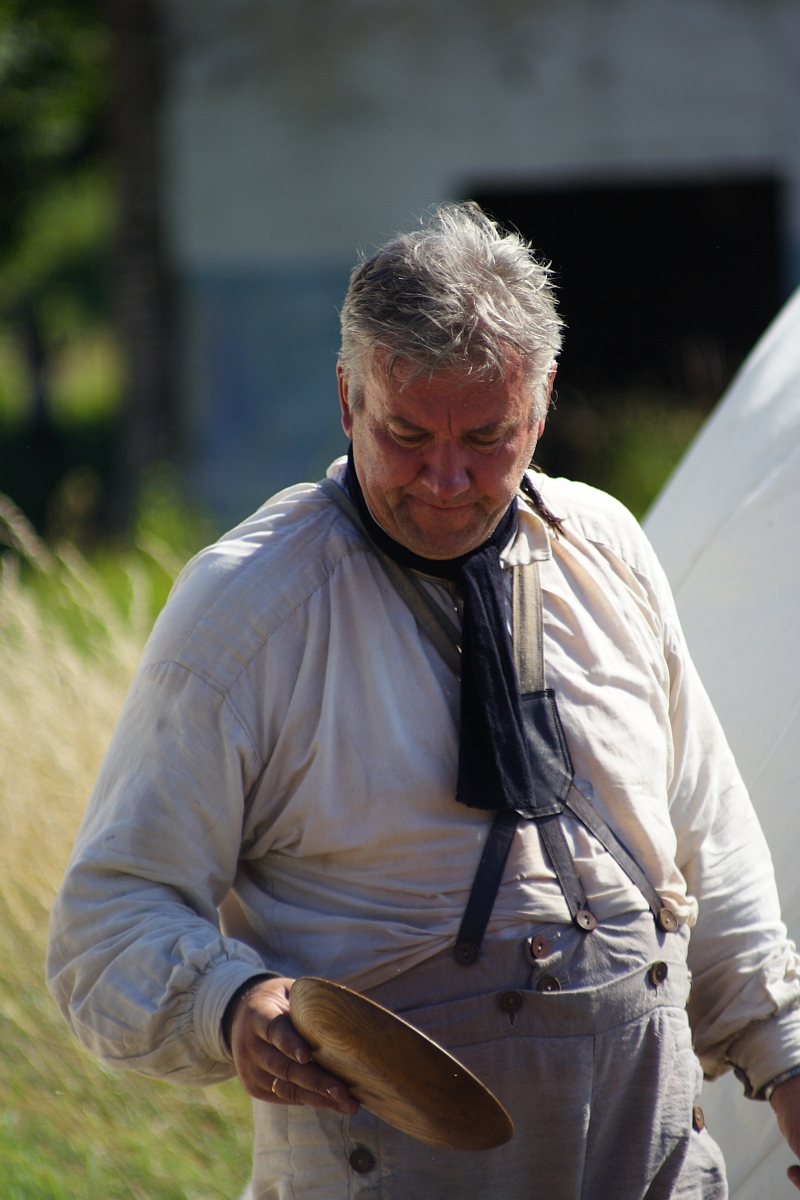 An older man with gray hair stands outdoors, wearing a light-colored long-sleeve shirt and gray overalls. He holds a wooden bowl in his right hand and has a dark scarf around his neck. The background is out of focus, showing some greenery and the vague outline of a building, reminiscent of Dunamunde 2010.