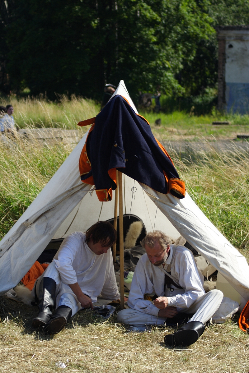 Two men dressed in white historical clothing sit under a white canvas tent pitched on grassy ground. The tent, draped with a dark cloak over the entrance, is set against a backdrop of tall grass, trees, and part of a building. Engaged in an activity, possibly writing or reading, they evoke the spirit of Dunamunde reenactments in 2010.