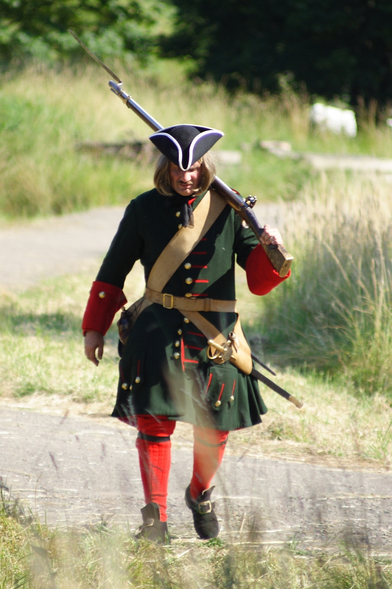 A person in a historical military uniform, reminiscent of the Battle of Dunamunde, walks outdoors on a sunny day. They wear a dark green coat with red and gold accents, a tricorn hat, red stockings, and black shoes. They carry a musket over their shoulder and a beige strap crosses their chest. Tall grass grows to either side of the path.