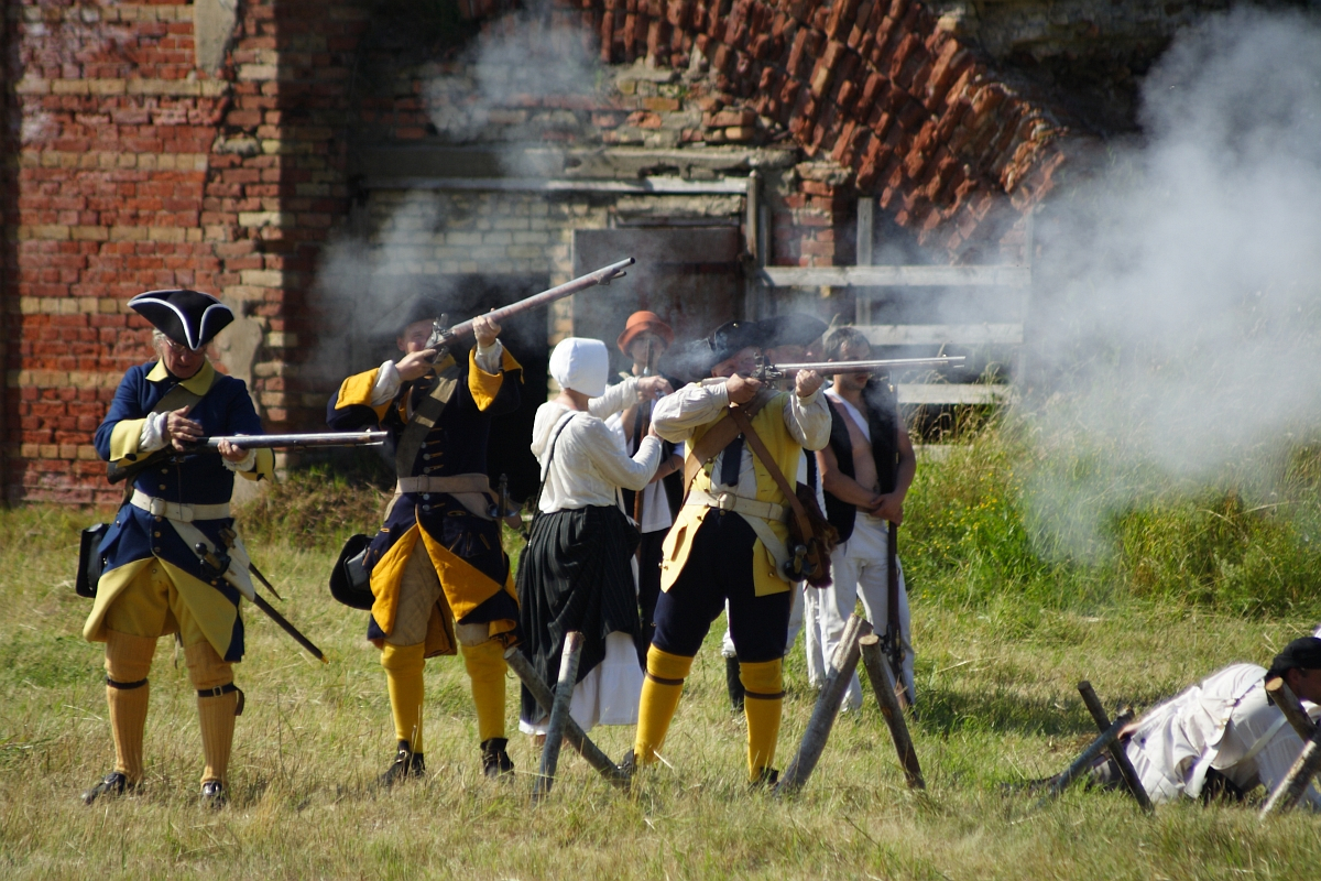 A group of historical reenactors, dressed in 18th-century military uniforms, are engaged in a mock battle at Dunamunde 2010. Four soldiers in yellow and blue uniforms are firing muskets amidst smoke. Behind them, a red brick building is partially visible, with grass and a wooden fence completing the scene.