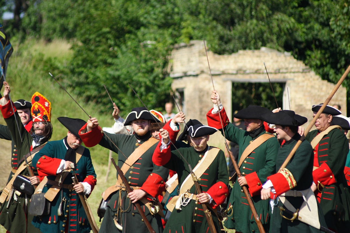 A group of costumed reenactors, dressed in historical military uniforms featuring black tricorn hats and green coats with red cuffs and lapels, raise their rifles in unison outdoors. Stood in front of an old stone structure with greenery in the background, they vividly bring to life Dunamunde 2010.