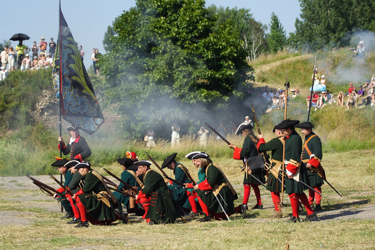 A group of historical reenactors dressed in green and red uniforms, complete with tricorn hats, participate in a battle reenactment reminiscent of the 2010 Dunamunde event. Some soldiers kneel while others stand, aiming long rifles. A large, ornate flag is held by one reenactor as spectators watch from a grassy hill.