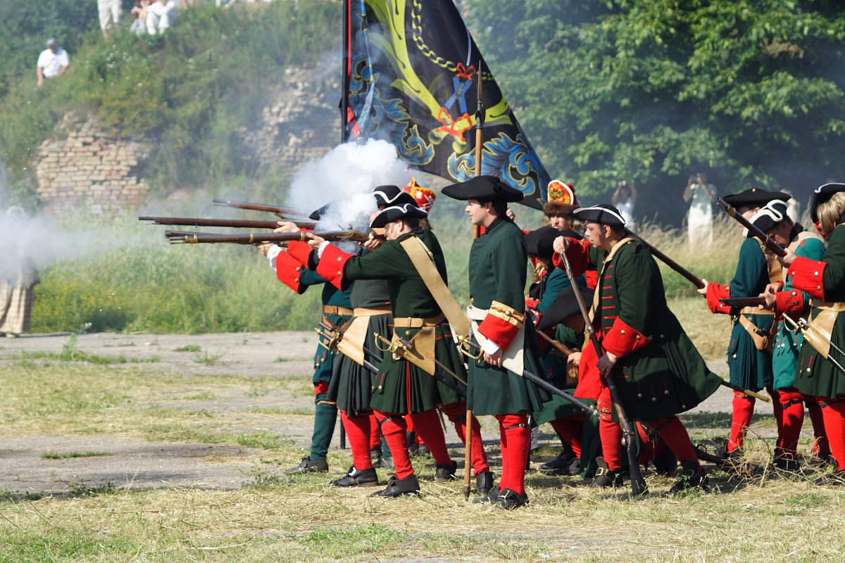 A group of historical reenactors in 18th-century military uniforms fire muskets during a staged battle at Dunamunde 2010. The soldiers wear dark green coats, red breeches, and black tricorn hats. Smoke emerges from the muskets. Trees and stone structures are visible in the background, along with a vibrant flag on display.