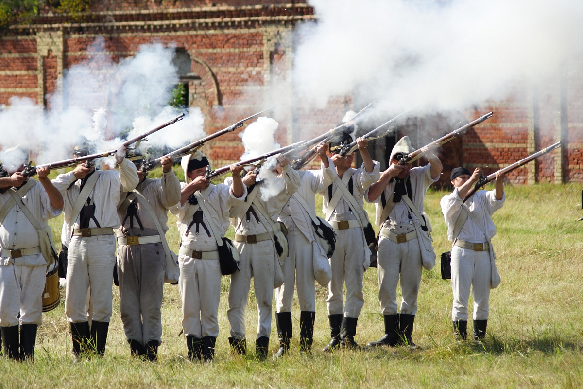 A group of people dressed in historical military uniforms stands in a row on a grassy field, firing old-fashioned muskets. Smoke from the gunfire fills the air. Behind them, there is a brick building with arched windows. The scene, reminiscent of Dunamunde 2010, appears to be a reenactment of a historical battle.