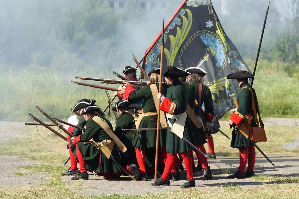 Historical reenactors at Dunamunde 2010 don 18th-century military uniforms, firing muskets and kneeling in formation. They wear dark green coats, red trousers, black tricorn hats, and carry long rifles. Smoke from the muskets fills the air as a complex black and yellow flag with a crest is held by one standing figure.