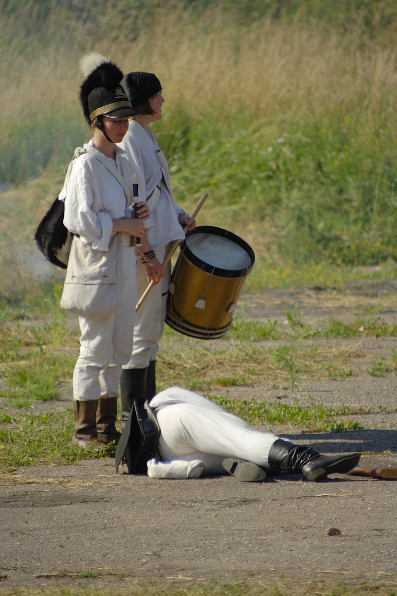 Two soldiers in white uniforms and black hats stand outdoors. One holds a drum, the other a flute. A person in a similar white uniform lies on the ground in front of them, appearing to have fallen, while the soldiers look on. The background features grass and foliage, evoking an eerie scene reminiscent of Dunamunde 2010.