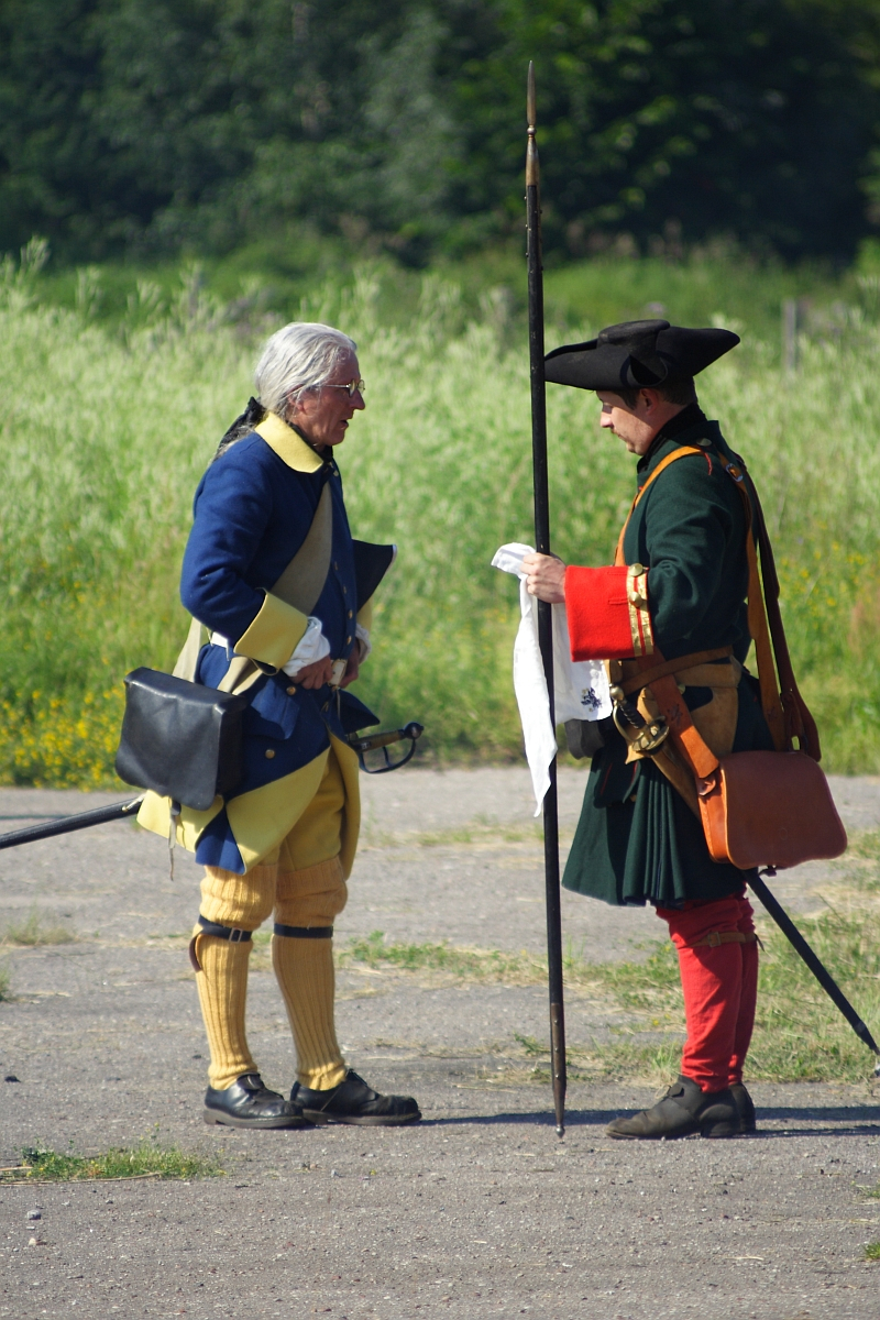 Two people dressed in historical 18th-century European military uniforms are engaged in conversation outdoors. One wears a blue and yellow uniform with a tricorne hat, while the other, donned in green and red, holds a spear in one hand and a white cloth marked "Dunamunde 2010" in the other. They stand on a dirt path with green foliage in the background.